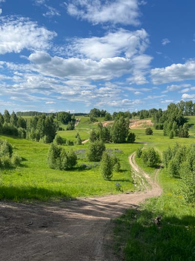 The image depicts a verdant, hilly landscape in Новочувашский, Russia, under a partly cloudy blue sky. The sky occupies the upper third of the frame, featuring scattered white cumulus clouds against a bright azure backdrop.

The midground and background are dominated by rolling green hills covered in lush grass and numerous deciduous trees of varying sizes, exhibiting bright green foliage. A prominent dirt track or path winds through the landscape, visible first in the mid-right and then extending further into the background, curving uphill towards the right. Another, more distant segment of this path zigzags up a large grassy hill on the far right. Several small, dark figures, identified as people, are visible walking on this distant uphill path. In the middle-right background, there is a distinct patch of exposed light brown/orange earth on a hillside, possibly indicating a natural erosion or a small excavation site. On the far left horizon, obscured by trees, two tall, thin crane-like structures are faintly visible, suggesting distant construction or industrial activity. Patches of purple wildflowers, likely lupines, are visible in the grassy areas, particularly on the left side of the midground.

The immediate foreground consists of a wide, rough dirt surface, partially covered by deep shadows cast from an unseen object to the left, indicating bright sunlight from the right. Visible roots protrude from the earth on the left edge of this foreground area. The dirt path is uneven, showing tire tracks or foot traffic marks. Green grass grows along the right edge of the foreground path.