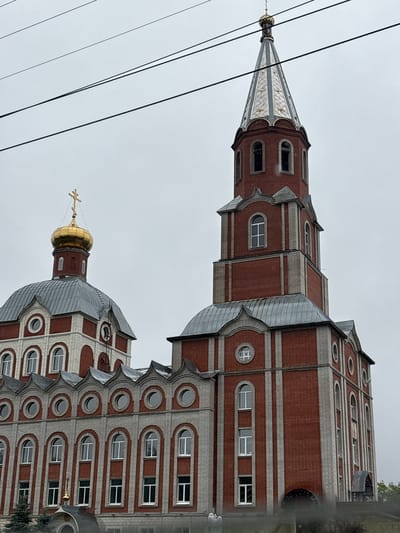 The image shows the Church of the Kazan Icon of the Mother of God in Krasnokamsk, Russia. The church is a red brick building with a tall, pointed steeple. The steeple is topped with a golden dome. To the left of the steeple is another dome, also golden and topped with a cross. The church has many arched windows and decorative elements. In the foreground, there are some power lines running across the sky. The sky is overcast and gray, suggesting a cloudy or rainy day. There are no people clearly visible in the image.