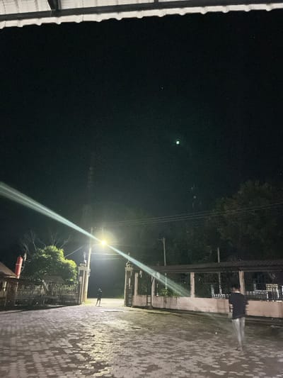 The image depicts a nighttime outdoor scene, viewed from a slightly elevated position. The orientation is portrait, but it appears to have been captured with the camera held horizontally and then rotated.

The sky occupies the upper left portion of the image and is dark, indicating night. Within this dark expanse, a prominent, bright green dot is visible, along with a much smaller, fainter white dot further to the left, possibly lens flares or distant light sources.

On the far left edge of the image, a textured, corrugated surface, likely a roof edge or building facade, is partially visible in white and grey tones.

The middle and right sections of the image are dominated by artificial light. A bright street light is positioned towards the lower right, emitting a powerful, elongated beam of light that streaks diagonally across the frame, creating a strong lens flare effect. This light source illuminates the immediate surroundings.

Below and to the right of the street light, a paved or tiled ground surface is visible, showing reflections or texture that might suggest it is wet or has a patterned design. In the background, beyond the paved area, there are outlines of trees and foliage, largely obscured by darkness but discernible against the ambient light.

Near the street light, a structure resembling a gate or entrance with pillars can be seen. A solitary human figure, mostly silhouetted against the bright light, stands near this gate or light source, facing away from the viewer. Utility poles and power lines are also visible in the vicinity of the street light and trees. The overall setting appears to be an urban or semi-urban area at night. No text is visible in the image.