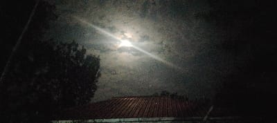 The image depicts a nocturnal scene in South Kasipul ward, Kenya. The upper portion of the frame is dominated by a dark, grainy sky with varying cloud density. A bright, circular light source, identified as the moon, is visible through partial cloud cover in the upper-mid section, emitting significant luminescence. A prominent diagonal light ray, consistent with an optical flare, extends from the moon's position towards the lower right. In the lower central and right portions of the frame, the reddish-brown corrugated roof of a structure is visible, with a gutter system discernible along its lower edge. To the left, a dense, dark silhouette of tree foliage or vegetation extends into the frame from the bottom left corner. No individuals, animals, or specific activities are discernible within the depicted environment.