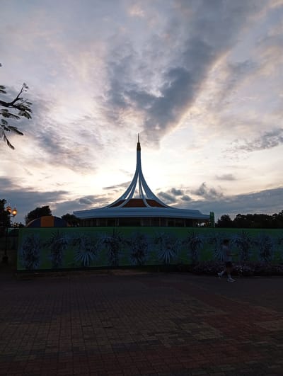 At dusk in Bang Kaeo Subdistrict, Thailand, a modern architectural structure with a distinctive spire stands silhouetted against a cloudy sky. The building features a circular base and an elevated, white roof with sharp, converging lines forming a central peak. In front of the building, a long banner with a green background displays a repeating pattern of stylized green palm leaves. To the right of the banner, a single person, appearing to be a male wearing a t-shirt and shorts, is captured in mid-stride on a patterned brick pathway. The pathway leads away from the viewer, toward the banner and the building. Scattered purple flowers are visible in a flowerbed between the pathway and the banner. The scene is illuminated by the soft, fading light of sunset, casting the foreground in shadow and highlighting the structure and the sky. To the left of the main structure, a tree branch with leaves extends into the frame, and two lampposts with visible lights are also present.