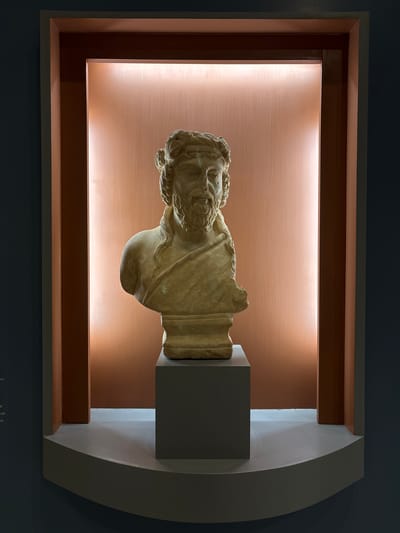A weathered, light-colored marble bust of a bearded male figure is centrally displayed within a recessed architectural niche. The bust depicts an individual with long, curly hair adorned with a wreath or headband, a prominent beard, and a draped garment over the left shoulder, terminating at the upper chest. The bust rests on a dark gray, rectangular pedestal, which is positioned on a light gray, curved base shelf that forms the bottom of the niche.
The interior walls of the niche are a reddish-brown color, illuminated by vertical strip lighting along the upper edges that casts a warm glow. The niche is framed by multiple layers of trim, featuring a darker reddish-brown inner frame followed by a lighter, almost beige-pink outer frame. This entire display is set into a larger wall painted a dark blue-gray or teal. Faint white text, indicative of an exhibit label, is visible on the dark wall to the left of the niche. This scene is located within a museum or exhibition space in the city of Drama, Greece.