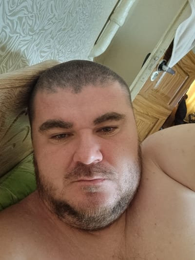 The image is a selfie taken by a man in what appears to be a bedroom in Romny, Ukraine. The man has short, dark hair and a beard. He is looking down and to the side. In the background, there is a patterned curtain, a door with a wooden finish, and a light-colored pipe. The man is lying down, with his shoulder visible in the right side of the frame. The overall lighting is soft, suggesting it might be daytime with natural light.