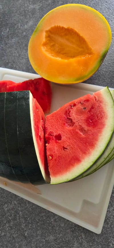 An overhead shot shows a whole cantaloupe and sliced watermelon on a white cutting board, which sits on a dark gray, speckled surface. The cantaloupe is a bright orange with a green rind, and it is cut in half, revealing the seedy interior. The watermelon is sliced into several wedges, with a dark green rind and bright red flesh. Some of the watermelon slices have been cut further into smaller pieces. The scene is brightly lit, with natural light casting soft shadows.