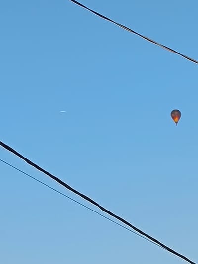 A hot air balloon, brightly lit with orange and yellow flames visible at its base, ascends into a clear blue sky. The balloon is positioned to the right of the frame. To the left of the balloon, a faint white contrail from an aircraft stretches horizontally across the sky. Two dark, thick power lines bisect the image diagonally, one from the upper left to the lower right, and another from the bottom left to the upper right, obscuring parts of the sky. The scene appears to be an outdoor setting, possibly in the city of Novonikolskoye, Russia, given the presence of power lines commonly found in urban and suburban environments. No people are visible in the image.