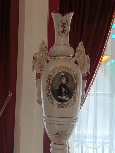 The image shows a tall, ornate white porcelain vase with gold trim and decorative elements. The vase features a portrait of a man in military attire holding a scroll within a circular, gilded frame. The top of the vase has a smaller decorative element depicting a figure on horseback. The vase is positioned indoors, with a rich red curtain in the background and sheer white curtains and a window visible to the right. A gold rope barrier with a pink knob is partially visible on the left. The scene is likely within a historical building or museum. This image was taken at the Kosava Palace complex in Kosava, Belarus, specifically within the museum exhibition.