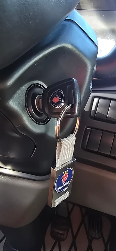 A close-up shot shows a truck's ignition and key. The key, with a Scania logo on it, is partially inserted into the ignition. A keychain with a larger Scania logo, a lion's head within a blue oval and the word "SCANIA" below it, hangs from the key. The ignition and surrounding dashboard are made of black plastic. To the right of the ignition, several dark, rectangular buttons are visible on the dashboard. The image is taken from a low angle, looking up towards the steering column and dashboard. The scene is indoors, likely within the cabin of a parked truck, with natural light filtering in from the top right, creating reflections on the plastic surfaces. The image was captured in Pedrola, Spain.