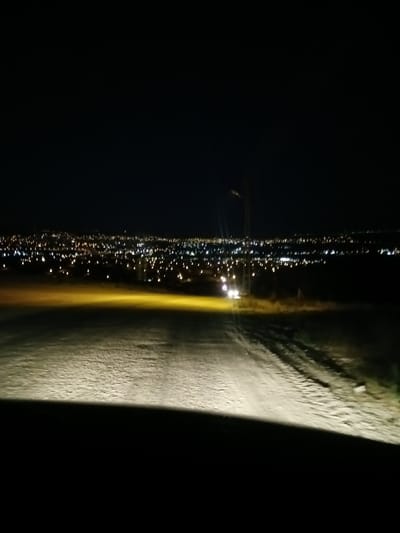 The image shows a dark landscape with a distant city illuminated by numerous lights. The foreground is a dirt road, likely illuminated by the headlights of a vehicle not fully visible, casting a bright, yellowish light. The road has visible tire tracks and a textured surface. Beyond the road, there is a field or undeveloped land. The middle ground features a prominent, thin vertical pole with a light fixture, a common sight in urban or suburban areas, and two bright white lights that appear to be from a vehicle driving on the road. The background is dominated by the sprawling city lights, which appear somewhat blurred, suggesting either distance, motion, or a shallow depth of field. The city lights stretch across the horizon, indicating a significant urban area. The sky above is completely black, devoid of stars or moon, indicating nighttime. The overall impression is that of a view from a vehicle approaching or overlooking a city at night from a slightly elevated position. The city is likely Bahşılı Belediyesi, Turkey, given the context.