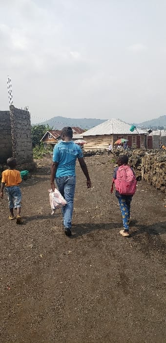 In Buzove, Democratic Republic of the Congo, three individuals walk away from the viewer on an unpaved, gravel-strewn dirt path. The central figure is an adult male wearing a light blue polo shirt and blue jeans, carrying a white plastic bag in his left hand. To his left, a child in an orange t-shirt and blue jeans walks ahead. To his right, a child wearing a pink backpack and patterned blue pants walks alongside. The path is bordered by a partially constructed stone block wall on the left and a low stone wall on the right. In the mid-ground, several single-story structures with various roof types are visible, including one with a light-colored corrugated metal roof and wooden walls. Distant mountains are visible under a light grey, overcast sky.
