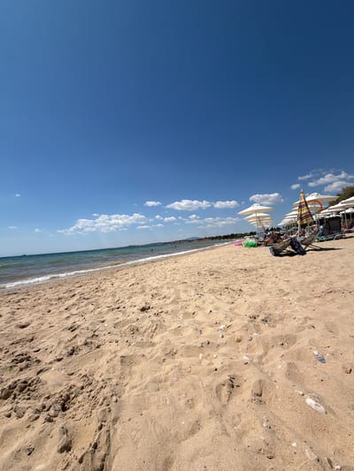 The image depicts a sunny day at a beach in Πόρτες, Greece. The foreground is dominated by a wide expanse of golden sand with footprints and small pebbles scattered across it. In the mid-ground, the gentle waves of the sea lap onto the shore. Further back, a row of white beach umbrellas provides shade for sunbathers relaxing on lounge chairs. A striped umbrella stands out amongst the white ones, adding a splash of color. The background shows a coastline with trees and some buildings, all under a clear blue sky with a few fluffy white clouds. The overall scene is one of a peaceful and inviting beach resort.