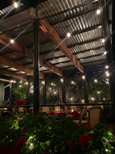This image displays an outdoor restaurant patio at night, located in Ridgefield, United States. The patio features a covered structure with a corrugated metal roof, supported by dark-colored vertical metal posts and horizontal wooden beams. Numerous string lights with exposed bulbs are strung across the roof structure and extend into the background, illuminating trees and shrubs. The patio is furnished with multiple wicker-backed chairs and circular red tables, some of which hold small illuminated objects, likely candles or lamps. In the immediate foreground, a planter contains dense green foliage interspersed with prominent red geraniums. One individual is partially visible seated at a table on the far left of the patio, their back mostly towards the viewer. No specific actions or interactions are discernible.