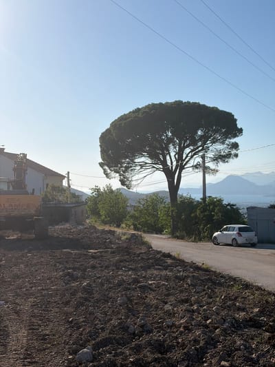 This detailed description outlines a sunny outdoor scene in Dobra Voda, Montenegro, featuring elements of construction, a road, vegetation, and a distant coastal landscape.

In the immediate foreground, a broad expanse of disturbed, dark brown earth, mixed with rocks and debris, covers the lower half of the frame. Faint tire tracks are visible on the left side of this excavated ground. To the left, a large yellow excavator is partially visible, its arm raised. The numbers "924" and the partial word "ALAMPEG" are discernible on its side. Directly behind the excavator, a white building with a distinctive red-tiled roof is visible.

Running diagonally from the lower right to the mid-left of the image is a narrow, paved asphalt road. On the right side of this road, a white compact hatchback car, identified as a Suzuki Swift, is parked facing away from the viewer. The car's rear license plate is partially visible, displaying "CG PV" followed by obscured characters. Adjacent to the car, a small, light-colored utility box or cabinet stands.

Dominating the central midground is a large, mature umbrella pine tree with a wide, flat-topped canopy of dense green foliage. A utility pole stands near its trunk, from which multiple power lines extend across the upper portion of the sky. Various other green trees and bushes are scattered throughout the midground, particularly behind the road and extending towards the background.

The background reveals a expansive body of water, likely the Adriatic Sea, stretching across the horizon. Beyond the water, several layers of mountains appear in varying shades of hazy blue, indicating distance and atmospheric perspective. The sky above is clear and a bright, light blue, with power lines traversing the upper frame. The scene is illuminated by bright sunlight, casting subtle shadows to the right, suggesting morning or late afternoon light. No individuals are visible within the frame.