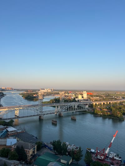 This is an aerial, wide-angle photograph of a city on a clear day. The sky is a pale blue, transitioning to a lighter shade near the horizon. The main subjects of the photo are two large bridges spanning a wide, calm river. The bridges are made of steel and appear to be older, truss-style structures with multiple spans. The river itself is a deep blue-gray, with gentle ripples on its surface.
On either side of the river, the city unfolds. The left bank shows a densely built urban area with apartment buildings and smaller structures. The right bank features a mix of industrial buildings, some with red roofs, and patches of green trees. In the distance, more city buildings and structures are visible, stretching towards the horizon. The overall impression is one of an established city with significant infrastructure.
In the foreground on the right side of the river, a port area is visible with several boats and a large red and white crane. The crane is positioned on a barge or ship, with its boom extended. The boats are of various sizes and appear to be moored. The architecture of the buildings on the left bank in the foreground suggests an older urban setting with brick and stucco facades. The scene is illuminated by what appears to be late afternoon or early evening sun, casting long shadows and bathing the scene in a warm light. There are no visible people in the image.
