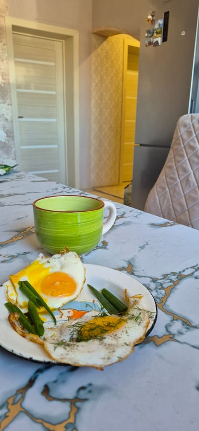The image depicts a breakfast scene on a marble-patterned table. In the foreground, a white plate holds two fried eggs, one with a fully cooked yolk and the other with a runny yolk, both garnished with chopped green onions and dill. To the left of the plate is a bright green mug with a white handle, filled with a light-colored liquid, likely coffee or tea. The background shows a kitchen environment. To the left, a closed door with horizontal glass panels is visible. In the center background, a yellow cabinet with a textured surface and a curved top is present. To the right, a portion of a gray refrigerator is seen, adorned with several small photos or magnets. A portion of a chair with a quilted, beige upholstery is also visible on the far right. The overall setting suggests a domestic kitchen or dining area. The scene is lit by natural light, casting shadows on the table. No individuals are present in the image. This scene could be located anywhere, including Yemelyanovo, Russia, but there are no specific visual cues to confirm this location.