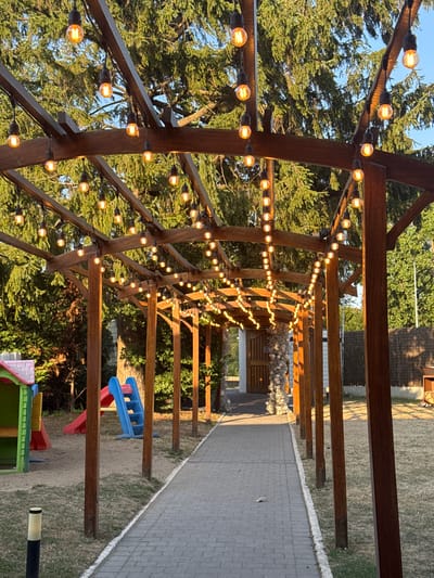 The image shows an outdoor pathway bordered by wooden posts and strung with incandescent lights, leading towards a closed wooden door. The pathway is paved with interlocking bricks and bordered by dry grass. To the left of the pathway, there is a children's play area with a small, multi-colored playhouse and a red and blue slide. The scene is set against a backdrop of lush green trees. The lighting suggests it is either early morning or late afternoon. The location is identified as Hem, France.