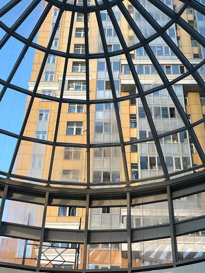 This media file captures a view through a modern glass and metal architectural structure, likely an atrium or covered walkway, looking out at an adjacent building. The structure features a grid of dark metal beams and clear glass panels, creating a geometric overlay on the scene beyond. The building viewed is a multi-story structure with a facade predominantly clad in a warm, golden-yellow material. Numerous windows, arranged in a regular pattern, punctuate the building's exterior. Some windows are clear, reflecting the sky and surrounding environment, while others appear to have blinds or internal coverings, creating variations in light and shadow. The reflections on the glass suggest it is daytime, with bright, possibly sunny, weather conditions, as indicated by the clear blue sky visible in some sections of the foreground and the strong reflections. There is no visible activity of people, nor any textual information. The perspective is from below, looking upwards and outwards, emphasizing the scale and modernity of the architecture. The overall impression is one of urban, contemporary design.