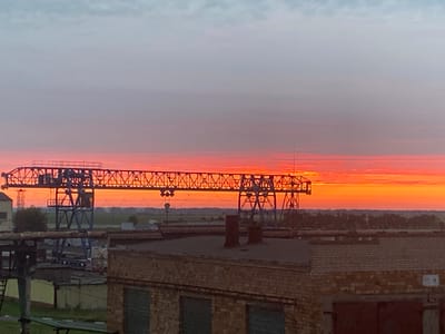 A wide shot captures an industrial landscape at sunrise, with the sky ablaze in vibrant oranges and pinks. In the foreground, the rooftops of brick buildings are visible. A large, blue metal gantry crane dominates the middle ground, its skeletal structure silhouetted against the colorful sky. Beyond the crane, a flat horizon stretches out, dotted with trees and fields, hinting at the rural surroundings of Краснадворскі сельскі Савет, Belarus. The scene is devoid of any human presence, emphasizing the industrial and natural elements.