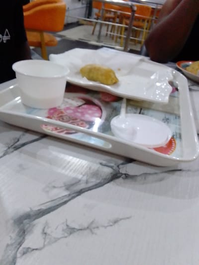 The image shows a white tray with a white cup, a white plate with a fried food item on it, and a white spoon. The tray has a pink and white design and is sitting on a white table. The table has a gray marble pattern. The location is in the city of Sapele, Nigeria, at the coordinates 5.936° N, 5.657° E.