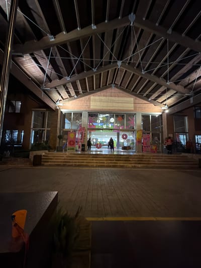 The image depicts an indoor scene, seemingly captured at night. The location appears to be a courtyard or atrium enclosed by buildings. The left side of the frame showcases an architectural structure with a wooden frame and what looks like a slatted or ribbed roof. This structure is held together by cables and beams.

The central area shows a multistory building front with several windows and decorative elements. Some windows have decorations resembling suns, flowers, and other artwork. There is also some signage visible.

The right side of the frame shows a stone wall or partition. There is greenery, possibly plants in a large container, partially visible. A partial view of a person's hand and what looks like a yellow object is seen in the lower right corner.

The environment suggests an urban area with a blend of architecture and decoration. The location is Punta Umbría, Spain.