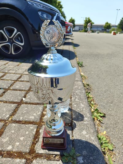 The image shows a silver trophy standing on a concrete sidewalk next to a black car. The trophy has a laurel wreath design on top with an emblem in the center. The base of the trophy is wooden and has text that reads "AVD - Club Orientierungs-Rallye." In the background, a person is walking across a paved area in front of a building. The scene appears to be outdoors on a sunny day. Based on the text on the trophy, it is likely located in Pirmasens, Germany, where the AVD (Automobile Club of Germany) may have a club that organizes orientation rallies.