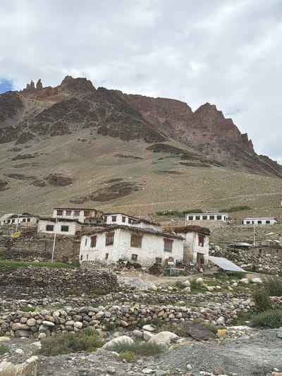 This is a vertical, eye-level, full shot of a remote village nestled at the base of a towering mountain range in Kurgiakh, India. The sky is overcast with heavy, gray clouds. The mountain is a dominant feature, with jagged, rocky peaks in shades of brown and gray, its slopes covered in sparse, dry vegetation and scree.
In the midground, a cluster of traditional white-washed houses with thatched roofs and wooden window frames is situated on a rocky slope. The houses are built with stone walls and plaster. Some of them appear to have solar panels installed. The foreground is a rugged terrain of loose rocks and pebbles, interspersed with dry grasses and small shrubs. There are no people or animals visible in the scene. The overall impression is one of a harsh, remote, and sparsely populated environment.