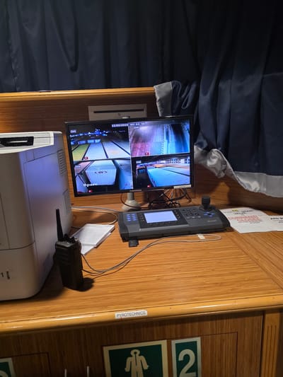 This is a photograph of a security monitoring station likely located in the city of Brindisi, Italy, possibly within a port or industrial facility given the context clues. The scene depicts a wooden desk surface with various electronic equipment.
On the left, a large, off-white Brother printer is partially visible. To its right, a black Motorola radio is positioned on the desk.
Dominating the center of the image is a multi-channel monitor displaying live feeds from four security cameras. The top left shows an overhead view of what appears to be a container yard with brightly lit containers. The top right camera feed shows a similar scene, possibly at a different angle or location, with more illumination. The bottom left camera displays a close-up of a white shipping container labeled "SPARE" and "R". The bottom right camera feed is partially obscured but appears to show a vehicle. The monitor also displays the date "22/06/2026" and time "00:43:48".
In front of the monitor is a control unit with a joystick, a touchscreen display, and a keypad, likely used for operating and navigating the camera feeds. A document, possibly an instruction manual or logbook, is placed beneath the radio.
To the right of the control unit, a sign is visible with red text stating "CURRENTLY CLOSED" and "HOSPITAL MAIN VALUE IS IN THE EQUIPMENT ROOM". Another sign with a green icon depicting a person walking and the number "2" is also visible towards the bottom right. A label on the desk's edge reads "PYROTECHNICS".
The background is dominated by dark blue curtains, which appear to be draped, suggesting an enclosed space like a control room or office. The overall environment appears to be functional and focused on security operations.