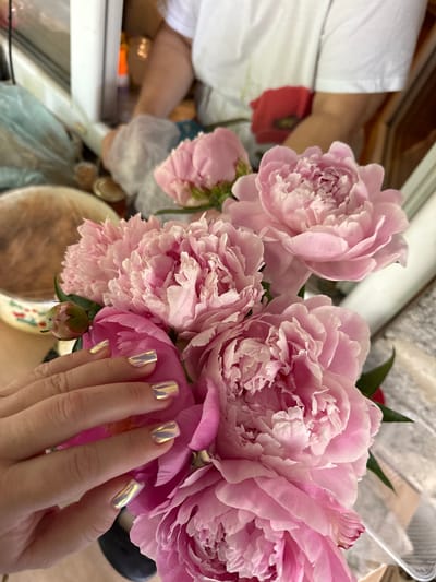 In Nizhne-Chufichevo, Russia, a close-up shot focuses on a bouquet of pink peonies, with a person's hand in the foreground. The hand, adorned with iridescent golden nail polish, gently touches one of the large, layered peony blooms. In the background, partially visible and out of focus, are elements suggesting an indoor setting with diffused lighting, possibly a kitchen or dining area, with a person wearing a white t-shirt in the far background. The overall scene captures a moment of appreciation for the floral arrangement.