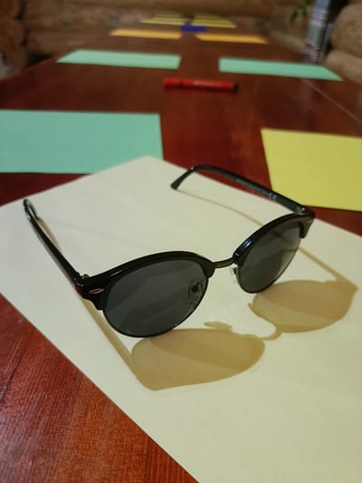 This is a medium shot, eye-level, indoor photograph of a pair of black-rimmed sunglasses with dark lenses, positioned in the foreground on a light beige surface. The sunglasses are angled with the left lens facing the viewer and are casting shadows on the beige surface. In the blurred background, a dark red wooden table is visible. Scattered on the table are several rectangular sheets of colored paper, including light green, yellow, and what appears to be a red marker or pen. The overall impression is that of a flat lay composition, likely set up for a visual project or activity. The location is not discernible from the image.