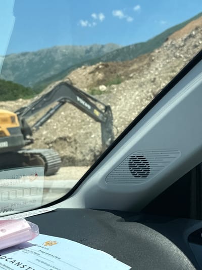This image appears to be taken from inside a vehicle, looking out through the windshield. The primary subjects are an excavator and a mountainous landscape. The excavator, a yellow Volvo, is actively engaged in what appears to be construction or earthmoving work on a rocky, uneven terrain. The background features a clear blue sky with a few white clouds, suggesting a sunny day, and rolling green hills that transition into a more rugged, rocky mountainside.

Within the vehicle, a grey speaker grille is visible on the interior paneling, suggesting the viewpoint is from the dashboard or A-pillar area. A document, likely official, is also partially visible on the right side of the frame. Some text from this document is legible in the OCR output, including "Crna Gora" (Montenegro), "Druga osnovna škola" (Second primary school), and potentially an address or identification number. The overall impression is a scene of ongoing work in a rural or semi-rural mountainous environment, viewed from the perspective of someone operating or riding in a vehicle. The time of day is likely daytime, given the bright sunlight and clear sky.