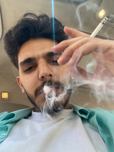 A man is smoking a cigarette indoors. He has dark hair and a beard, and is wearing a white shirt with a light blue jacket. The location appears to be inside a car, based on the overhead interior lights and roof. The city of Ma'loula, Syria, is not discernable from the visual information provided.