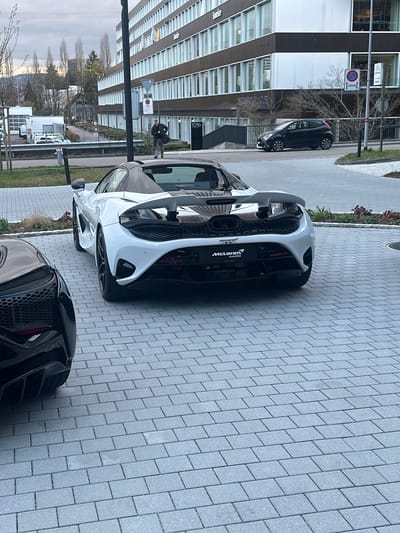 The image shows a McLaren supercar parked on a paved area in Glattbrugg, Switzerland. The car is positioned near a building with glass windows, and a second black car is partially visible in the foreground. People are present in the background, and street signs are visible.