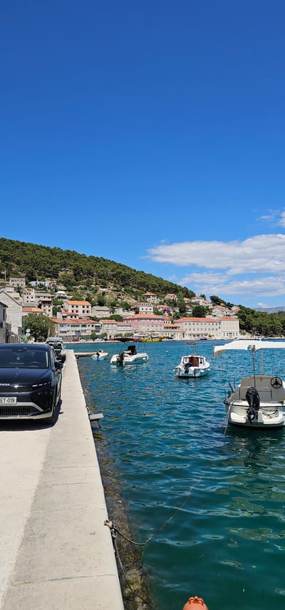 The image depicts a coastal scene in Općina Pučišća, Croatia, under a clear blue sky with scattered clouds. In the foreground, a black car is parked alongside a concrete walkway that borders the blue-green sea. Several small boats are moored in the water, some with canopies. Across the water, a town is built into a hillside, characterized by buildings with reddish-orange tiled roofs, interspersed with green trees and vegetation. The architecture suggests a Mediterranean style. The overall impression is a serene, sunny day in a picturesque seaside town.