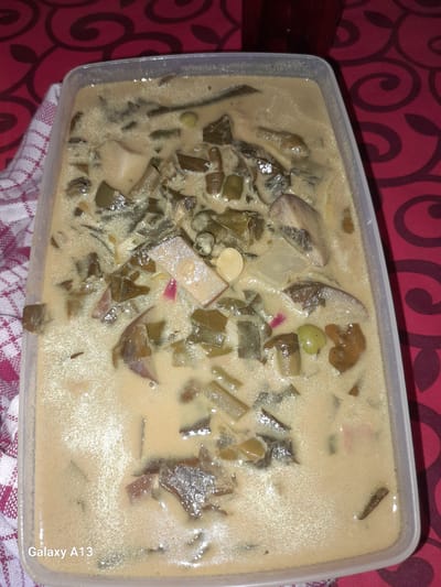 A close-up, overhead shot shows a rectangular plastic container filled with a creamy, light brown stew. The stew contains various ingredients, including chopped green vegetables that appear to be cooked leafy greens, diced white root vegetables, and some reddish-purple slivers that could be onion or other vegetables. The surface of the stew is glossy, indicating a liquid base, likely coconut milk or a similar creamy sauce. The container is placed on a red tablecloth with a swirling pattern. A white and red checkered cloth is draped on the left side of the container. A dark red beverage in a glass is partially visible in the upper right corner. The "Galaxy A13" watermark is present in the lower left corner. The scene is indoors, on a table.
