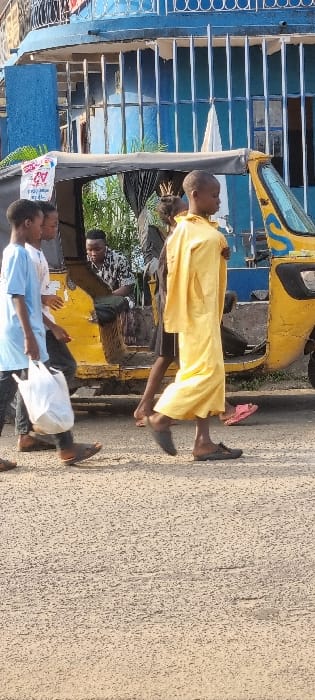 A motorized rickshaw sits parked by the side of the road with three passengers inside. One passenger exits the rickshaw, clad in a yellow robe and dark shoes. Beside him, another passenger wears a white shirt and carries a white plastic bag. The rickshaw has a yellow and blue color scheme and sits beside a blue building with a barred window. A textured, light-colored road surface stretches out to the side of the rickshaw.