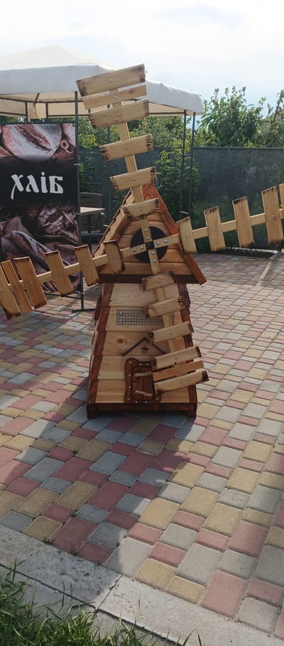 This is a medium shot, outdoor photograph of a wooden windmill structure placed on a patterned paver patio. The windmill is constructed from light-colored wood with dark-stained edges. It has four rotating blades made of wooden planks attached to a central black disc. The windmill has a peaked roof, a small door with a window-like grate above it, and decorative wooden elements.
In the background, a large banner with the Cyrillic word "Хлiб" (meaning "bread") is visible, along with an image of loaves of bread. Behind the banner and to the right of the windmill, there is a dark fence and green foliage. A white canopy structure is visible in the upper left quadrant of the image. The ground is paved with interlocking rectangular pavers in shades of red, yellow, and gray. A small patch of green grass is in the bottom left corner of the image. The lighting suggests it is daytime, with shadows cast on the ground.
The image was taken in the city of Ploske, Ukraine. There are no people visible in the photograph.