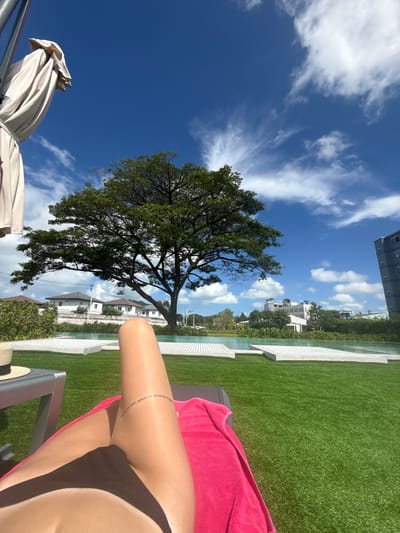 The image is a point-of-view shot from someone relaxing on a lounge chair by a pool on a sunny day in Ratsada, Thailand. The foreground shows a person's legs and torso, wearing a dark bikini bottom and a pink towel draped over their legs. A thin tattoo is visible on the upper thigh. To the left, a portion of a lounge chair and a straw hat are visible. Beyond the lounge chair is a bright green lawn with a large, mature tree casting shadows. In the mid-ground, a swimming pool with white tiling and clear water is present. Behind the pool, houses and buildings are visible, suggesting a residential or resort area. The background features a clear blue sky with scattered white clouds and a tall, dark building on the right side of the frame. The overall scene depicts a leisure setting with a tropical ambiance.