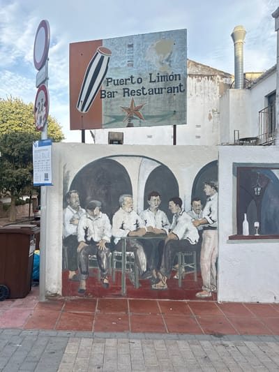 This image depicts a building wall in Calella De Palafrugell, Spain, featuring a large mural and an overhead sign.
Above the wall, a weathered rectangular sign for "Puerto Limón Bar Restaurant" is displayed. It features a striped, elongated drum graphic on an orange panel and a reddish-orange five-pointed star below the text, set against a light blue background.
The wall below the sign hosts a large mural. This mural portrays six men in what appears to be an interior bar or tavern setting with three prominent dark arches. Five men are seated around a dark rectangular table, all wearing white collared shirts and dark trousers; one seated man has a dark flat cap. A sixth man stands to the right of the table, holding a small glass, engaging in conversation with a seated man. The mural's floor is painted in reddish-brown tiles. To the immediate right of the main group, a painted window on the mural shows a wine bottle and a glass containing red wine on a sill, with an interior view of a hanging lantern beyond.
To the left of the mural, a vertical pole stands, bearing three signs: a red circular no-entry sign, a red circular sign with "2T" and "27" possibly indicating a vehicle restriction or towing information, and a white rectangular "TARIFES" sign listing prices. At the base of this pole, on the sidewalk, are two brown wheeled refuse bins and a blue plastic bag.
The ground in the immediate foreground consists of grey paver stones, transitioning to reddish-brown square tiles directly in front of the mural wall. The background above the mural and behind the sign reveals parts of an older, light-colored building, including an exposed metal vent pipe on an upper level and a small balcony with a metal railing. The scene is illuminated by daylight under a clear sky.