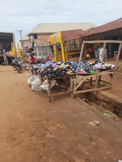 This image depicts an outdoor market scene in Bokkos, Nigeria. In the foreground, a wooden table is laden with a large assortment of colorful flip-flops and sandals. Beneath the table, several large, light-colored sacks are piled up. To the left of the table, a motorcycle is parked. Behind the market stall, a yellow structure, possibly a kiosk or booth, is visible, along with various shops with corrugated metal roofs. A truck with a large load is parked to the far left. Several people are present in the scene, some appearing to be vendors and others customers. The ground is unpaved and appears to be made of reddish-brown dirt. A narrow, open drainage ditch runs along the right side of the image. Overhead, power lines are visible against a cloudy sky.