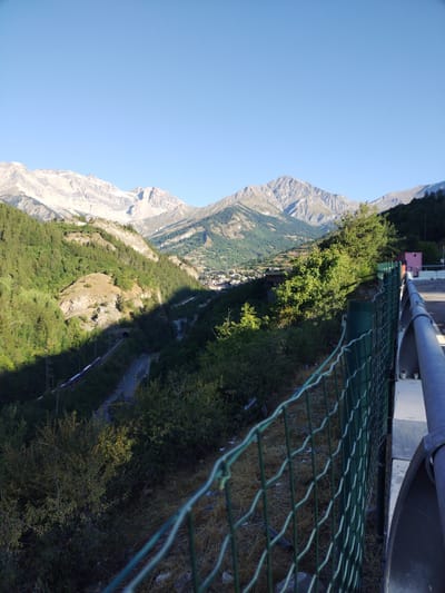 The image depicts a scenic mountain landscape with a village nestled in the valley. The foreground features a green metal fence and a grey metal railing, suggesting a viewpoint or a roadside. Beyond the fence, a train track winds its way through a lush green valley, disappearing into a tunnel on the left side of the image. The valley is densely populated with trees, and a small village with buildings is visible in the distance. Majestic, snow-capped mountains form the backdrop, their rugged peaks reaching towards a clear blue sky. The lighting suggests it is daytime, with shadows cast across the landscape, indicating the sun is at an angle. The overall scene captures the natural beauty of the mountainous region, with the human settlement integrated into the environment. This view is consistent with locations in or around Bardonecchia, Italy, a town known for its Alpine setting and railway infrastructure.