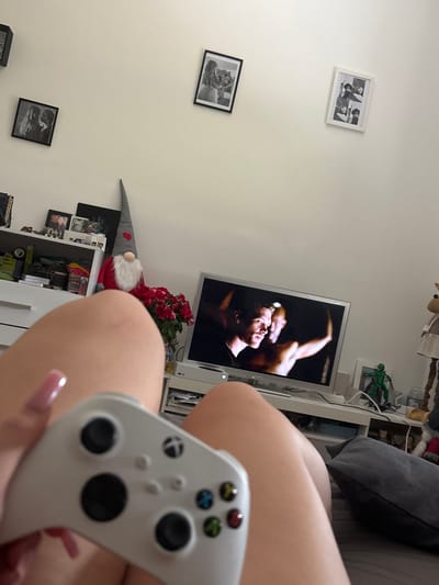 The image shows a person holding a white video game controller, with their legs in the foreground. In the background, a television screen displays a scene from a movie or show. A shelf unit is visible, containing various items including framed photographs, a gnome figurine, and a bouquet of red roses. The room appears to be a bedroom or living area, with a bed and a pillow. The setting does not provide any specific information about the city of Hausach, Germany.