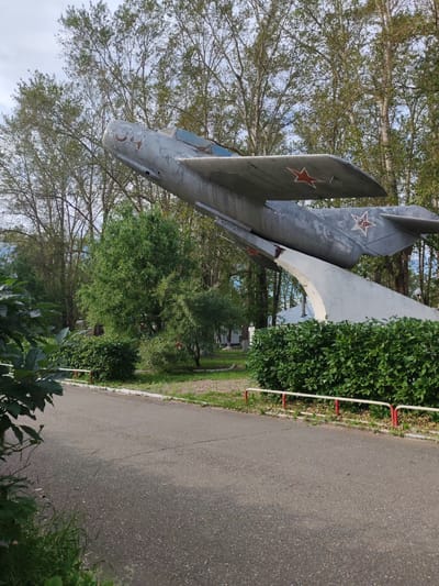 This media file shows a weathered, gray fighter jet monument displayed outdoors in a park-like setting. The jet is tilted upwards, supported by a concrete structure. Red stars are visible on the wings and tail of the aircraft. The number "04" is painted on the front of the fuselage.

The scene is surrounded by lush green trees and bushes, suggesting a natural environment. A paved path runs in front of the monument, bordered by a low fence with red and white posts. The ground is covered in grass and some patches of pavement. In the background, glimpses of buildings can be seen through the trees.

The lighting and the presence of shadows suggest it is daytime, likely overcast or with dappled sunlight filtering through the trees. There is no discernible activity taking place; the scene appears to be a static display. The overall impression is one of a memorial or historical exhibit, situated in a public outdoor space in Сокол, Russia. The monument itself shows signs of age, with chipped paint and wear, indicating it has been on display for some time.