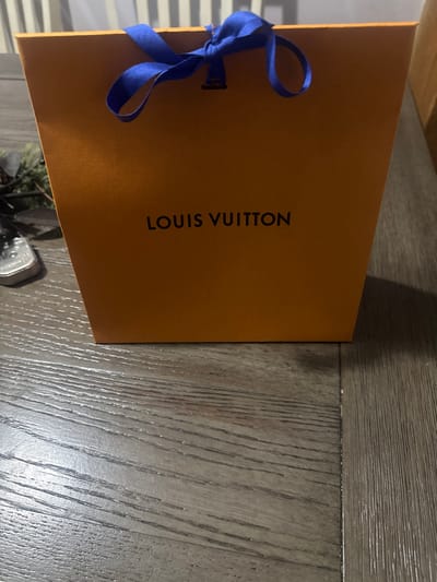 The image displays a vibrant orange Louis Vuitton shopping bag, positioned horizontally across the frame with its printed side facing the viewer. The brand name "LOUIS VUITTON" is visible in black capital letters, printed vertically along the length of the bag. A bright blue ribbon is tied into a bow, situated towards the top-left edge of the bag. The bag rests on a textured wooden surface, characterized by a distinct wood grain and horizontal plank lines, which covers the majority of the background and foreground to the right and bottom of the bag. On the far left, a blurred background element with horizontal striations suggests either a window with blinds or a wall paneling. A small, dark, indistinct object, possibly a potted plant or piece of furniture, is partially visible in the bottom-right corner. The scene is illuminated by soft, ambient indoor lighting, casting subtle shadows from the bag. The photograph is a still life, primarily showcasing the luxury brand's packaging.