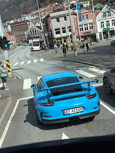 This image presents an overhead view of a bustling street scene in Bergen, Norway, bathed in bright sunshine. The central subject is a striking light blue Porsche GT3RS sports car, seen from the rear, stopped on a road with pedestrian crossings. Its Norwegian license plate, "BT 42549," includes the text "Porsche Center Bergen," confirming the location.
The scene depicts an active urban environment with various people and vehicles. Pedestrians are scattered along the sidewalks, some standing, others walking. A dark-colored car is partially visible in front of the Porsche. Further down the road, a white bus displaying "ASKO" on its side is seen, along with a "30" speed limit sign. A traffic light in the foreground is green.
Buildings of different architectural styles line the street. Notable storefronts include "Specsavers Optikk" (Optics) and another sign indicating "BRILLER KONTAKTLINSER" (Glasses Contact Lenses). A shop named "Hårtjen Frisør" (Hairdresser) is also visible. A blue banner with the text "OCEAN WEEK" can be seen. One person in the foreground is wearing shorts with "ASEGLA" printed on them. The sharp shadows cast by objects and people indicate clear weather and bright daylight, likely midday to early afternoon. The image is captured from an elevated perspective, looking down onto the street below.