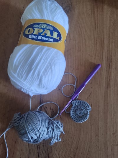 This is a top-down, medium shot of crochet supplies on a wooden surface. The main subjects are two balls of yarn, a crochet hook, and a small crocheted circle.

One ball of yarn is large and white, with a yellow and blue label that reads "OPAL Dört Mevsim". The other ball of yarn is smaller and grey, with loose strands of yarn trailing from it. A purple crochet hook is partially visible, attached to the grey yarn and inserting into the grey crocheted circle. The crocheted circle is small and round, appearing to be in the process of being made, with visible stitches. The wooden surface has a prominent grain pattern.

The scene suggests a hobby or craft activity is in progress, likely knitting or crocheting. The lighting suggests it is daytime. There are no people visible. The location is specified as Batken, Kyrgyzstan, but there are no specific visual cues in the image to confirm this.