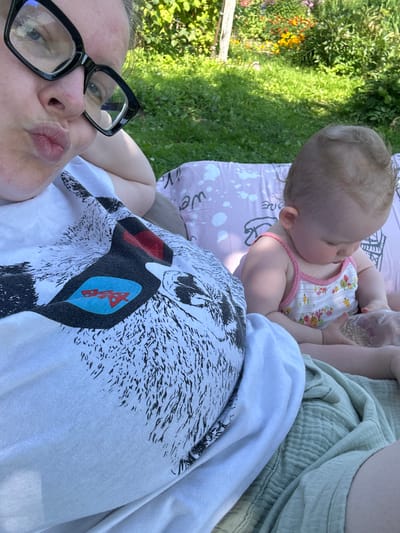 A woman with glasses is taking a selfie outdoors in what appears to be a yard with green grass and flowers in the background. She is wearing a white t-shirt with a graphic print and is puckering her lips. Next to her, a baby in a floral tank top is sitting and playing with a clear plastic bag filled with small objects. The location is not specified in Vereiki, Russia.