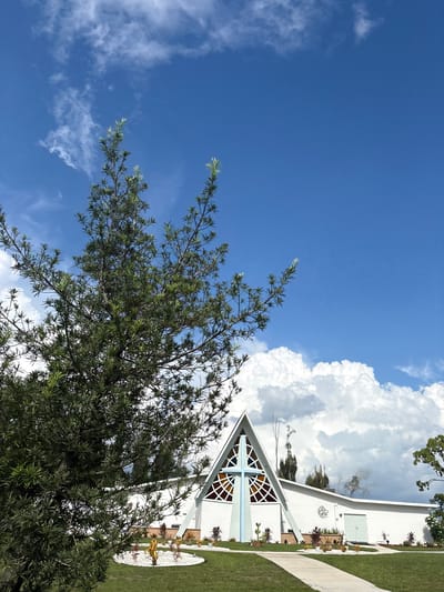 The image shows a modern white church building with a triangular roof and stained-glass windows forming a cross shape. A light blue cross is prominently displayed in front of the building. The church is surrounded by a manicured lawn and a pathway. In the foreground, a large evergreen tree with delicate branches partially obscures the view of the building. The sky is bright blue with fluffy white clouds, suggesting a sunny day. The location is identified as the city of North Port, United States of America. There are no people visible in the image.