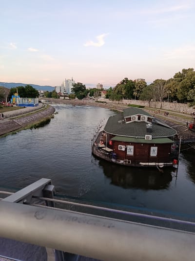A wide shot captured from a bridge overlooks the Nišava River in Niš, Serbia. The river flows through an urban environment with buildings and trees lining its banks. In the foreground, a section of the bridge railing with cables is visible. The midground features a distinctive, oval-shaped wooden houseboat with a green roof, appearing to be a restaurant or bar, with people seated at tables on its deck. The river's surface is dark and reflects the sky and the houseboat. The background shows a cityscape with modern buildings, a stone-walled embankment, trees, and distant hills under a sky with soft clouds, indicating either early morning or late evening light. On the left bank, an inflatable amusement structure and people are visible near the river. The scene depicts a lively, urban waterfront setting.