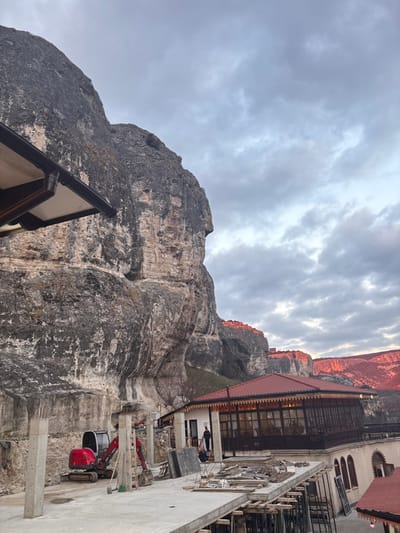 The image displays a construction site at the Holy Dormition Cave Monastery (Uspensky Monastery) in Bakhchysarai, Ukraine, situated directly against a prominent, weathered grey and tan limestone cliff. The sky is uniformly overcast with grey clouds. In the mid-ground, a concrete slab is under construction, supported by wooden scaffolding and several unfinished concrete pillars. A compact red excavator with a grey cab and bucket arm is parked on disturbed earth at the base of the cliff, positioned behind the concrete work. To the right, an existing two-story building features a red gabled roof, light-colored walls, and an enclosed upper-level balcony with dark wooden latticework and multi-paned windows. Adjacent to this structure, a simpler, single-story light-colored building has two individuals dressed in dark attire standing near its entrance. In the background, distant cliffs and mountains are bathed in a distinct reddish-orange light, indicating a low sun angle. The scene depicts ongoing construction or renovation activity integrated into a historical mountainous landscape.
