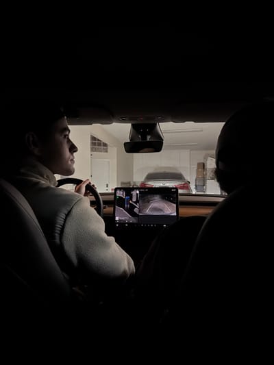 The image shows a person sitting in the driver's seat of a Tesla, possibly in a garage. The person is holding the steering wheel and looking upwards towards a tablet mounted on the dashboard, which displays a visual interface that may be related to self-driving or parking assist features. The garage contains another vehicle and some storage boxes. Based on visual characteristics, the location is likely Santa Ana, United States.