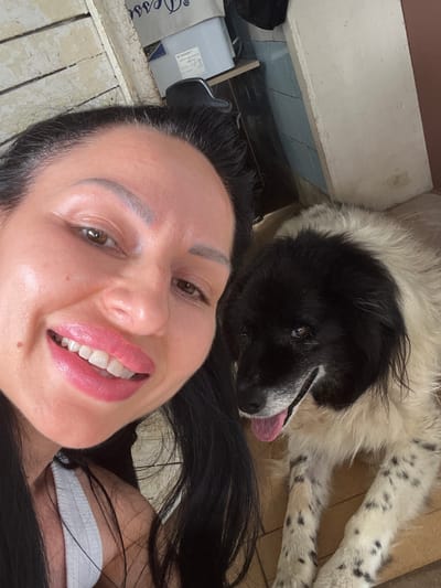 A selfie of a woman and a dog is captured in an indoor setting. The woman, with long dark hair, smiles broadly, her lips glossy and pink. She wears a white top. Beside her, a large dog with black and white fur lies down, its tongue hanging out. The background shows a tiled wall and what appears to be a weathered wooden structure. The lighting suggests it is daytime. The location is specified as Ohrid, Bulgaria, though no specific landmarks or city features are visible to confirm this. The scene is informal, suggesting a personal moment between the woman and her pet.